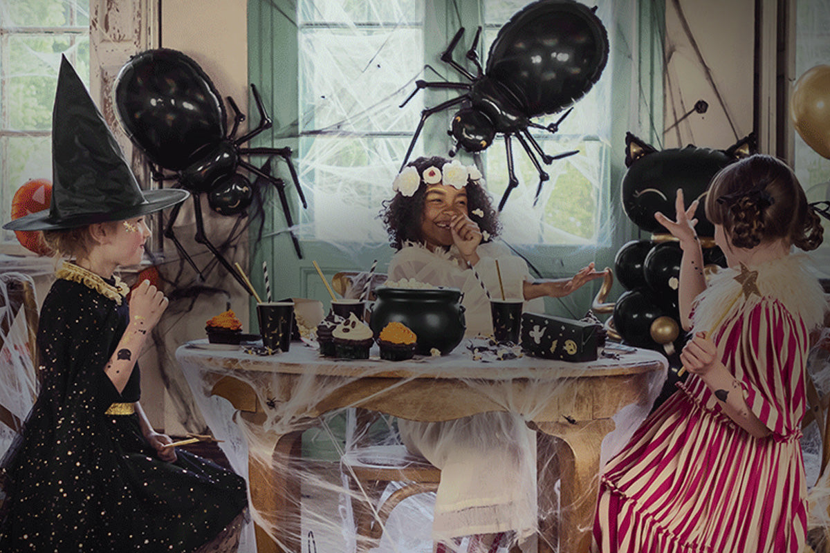 Three children in Halloween costumes sitting around a table with large spiders and balloons in the background.