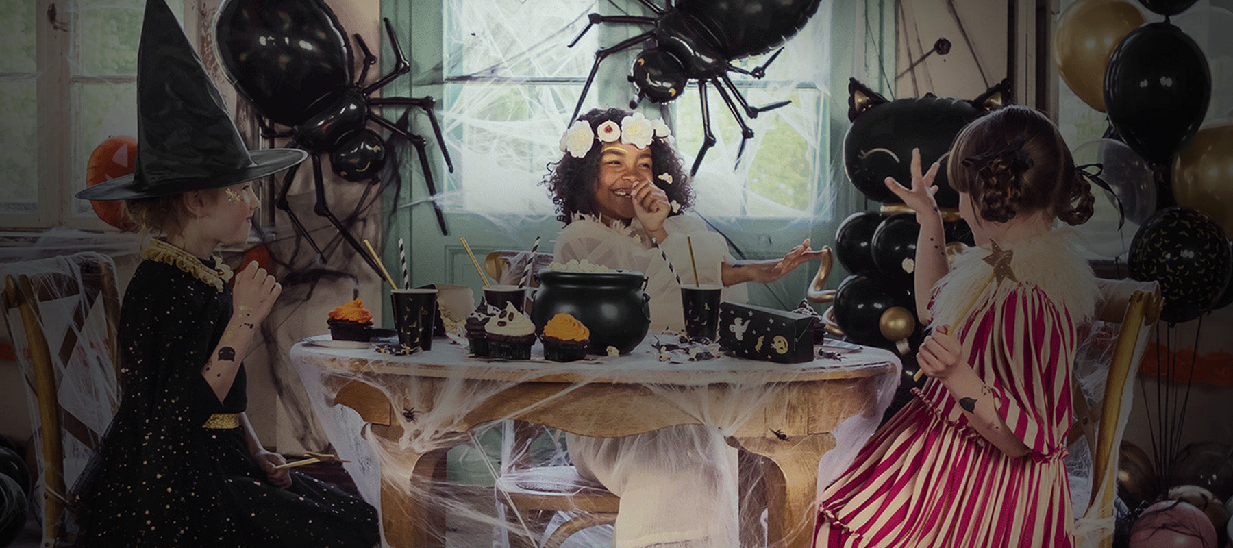 Three children in Halloween costumes around a table with decorations, including spiders and pumpkins.