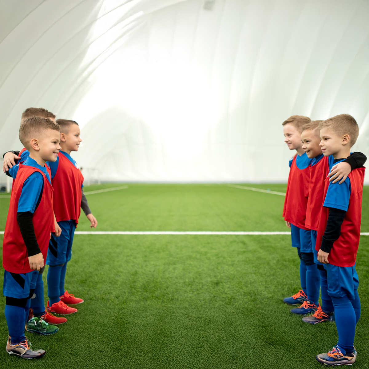 Children in red and blue sports uniforms standing on a green indoor soccer field.