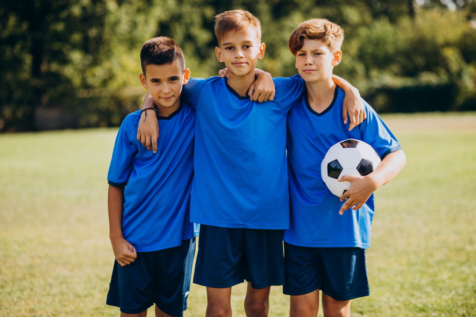 Three boys in blue soccer jerseys standing on a grassy field with a soccer ball.
