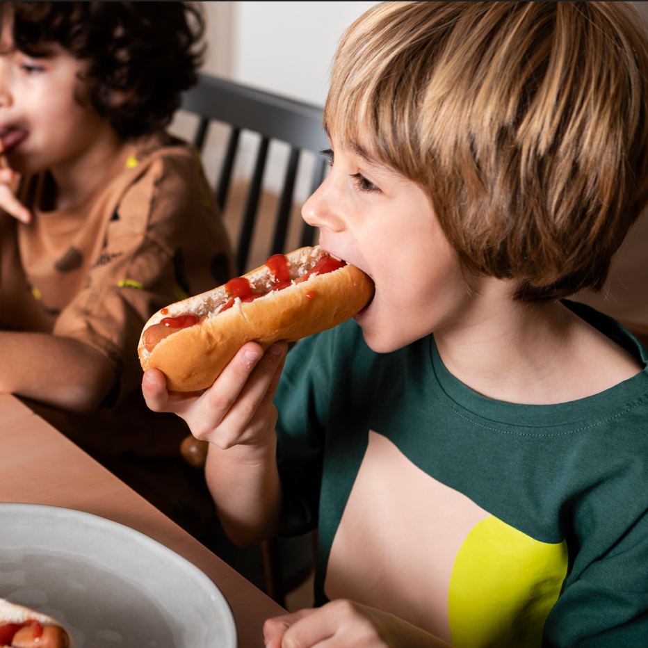 Child eating a hot dog at a table with another child in the background.