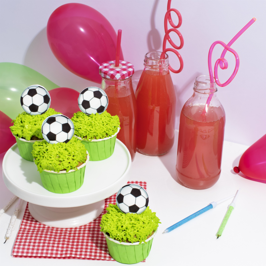 Cupcakes with soccer ball toppers on a cake stand with juice bottles and balloons in the background.