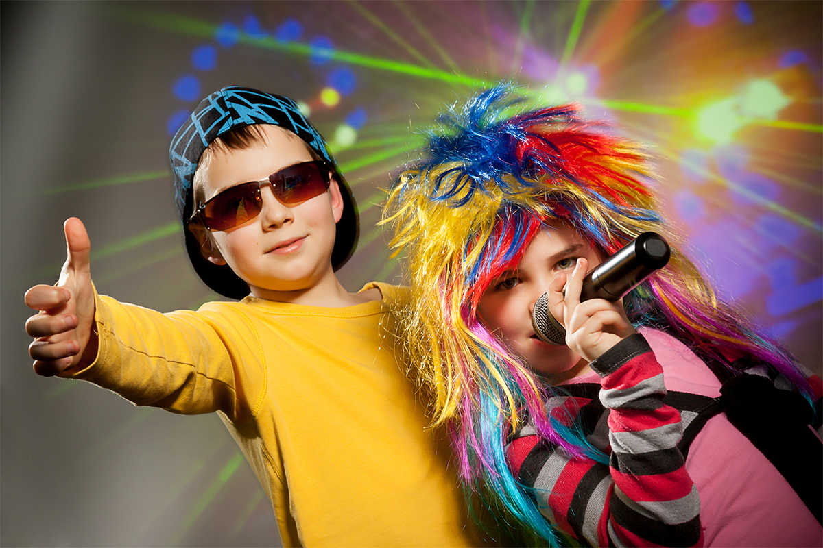 Two children in colorful costumes with a microphone and sunglasses, performing on stage.