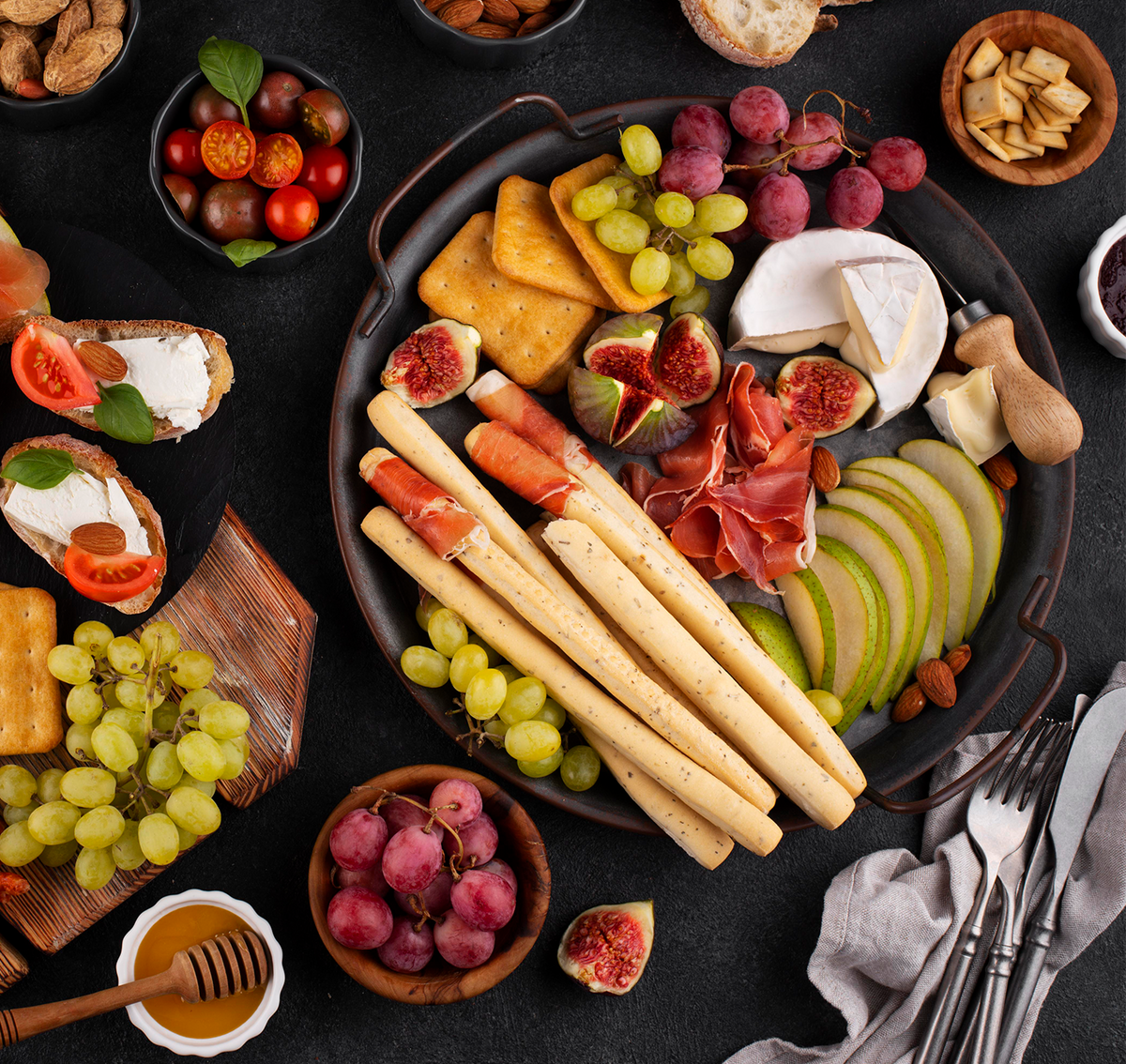 Assorted platter of cheese, fruits, and bread on a dark surface with utensils.