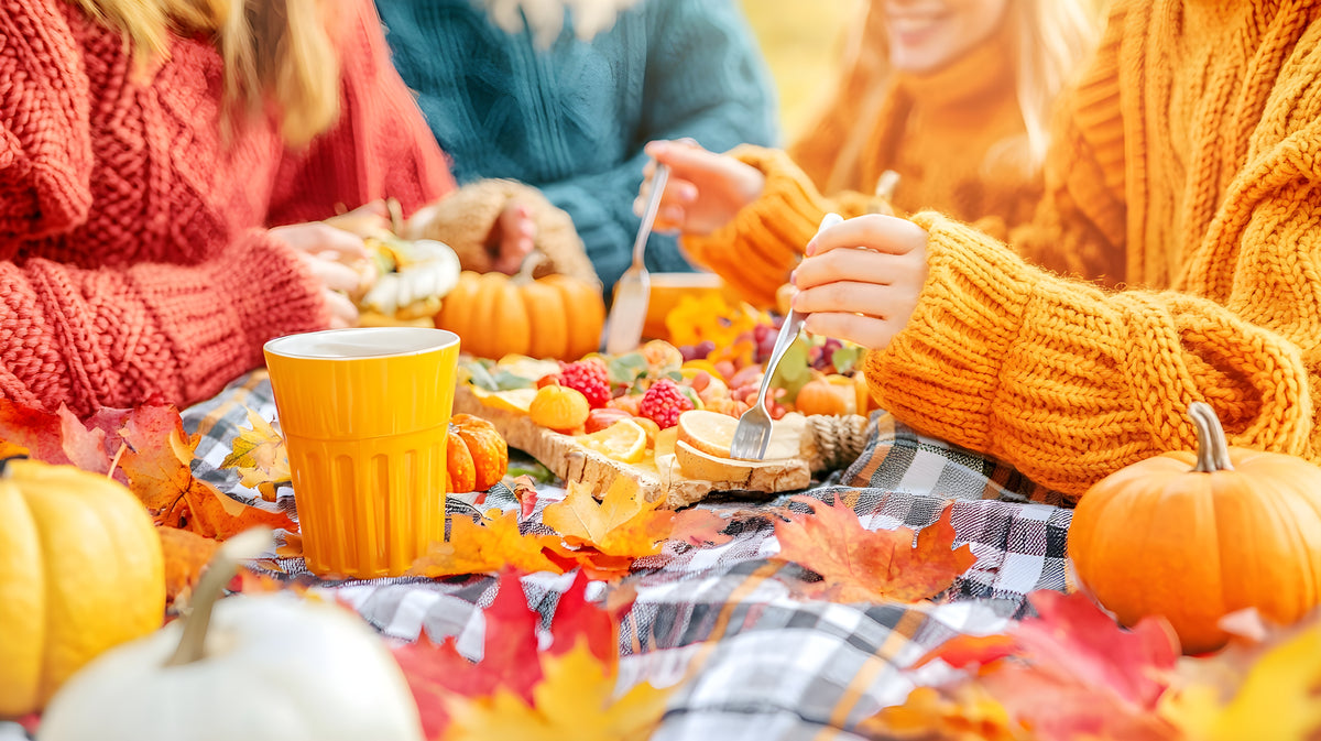 People enjoying a meal outdoors with pumpkins and autumn leaves on a checkered blanket.