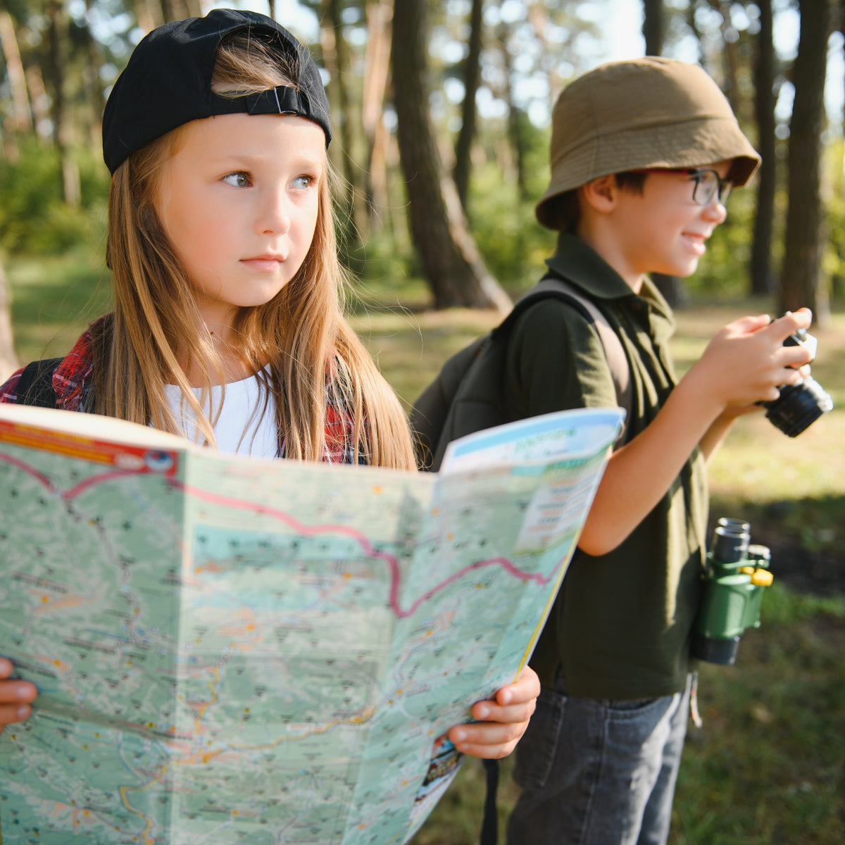 Two children outdoors, one holding a map and the other with a camera, in a forest setting.