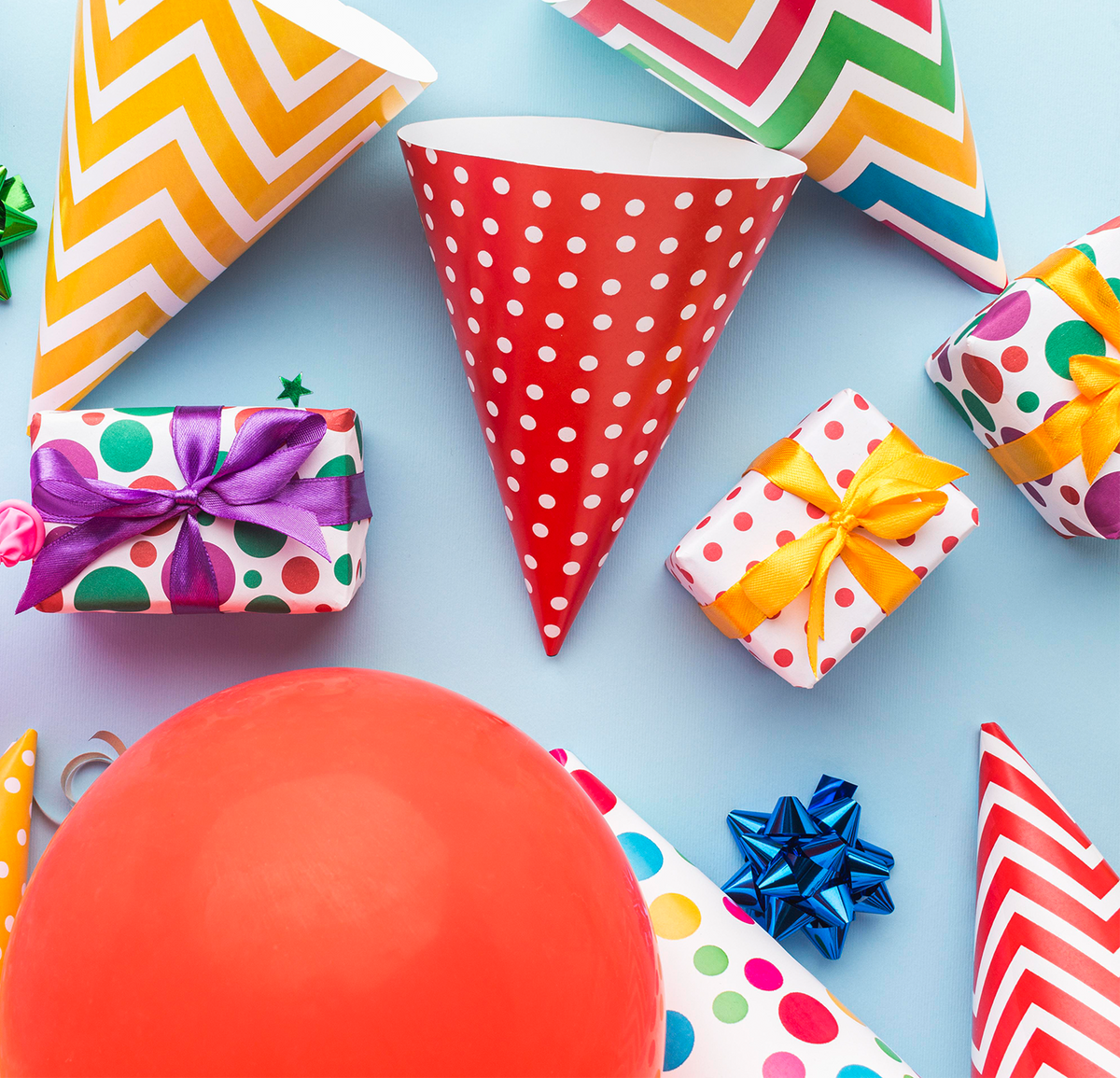 Colorful party supplies including balloons, gift boxes, and cones on a light blue background